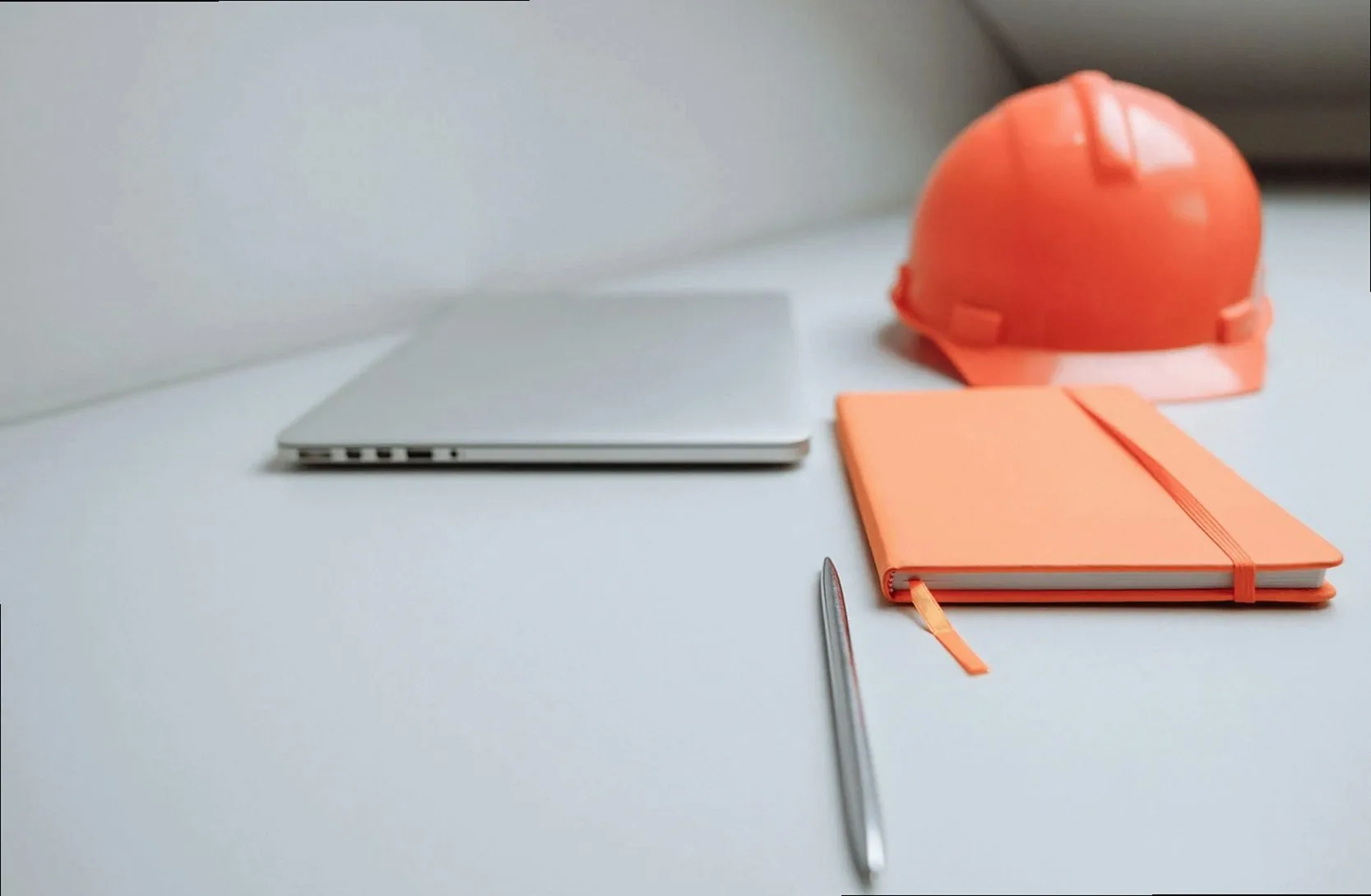 A minimalist office setup featuring a laptop, orange notebook, and safety hard hat.